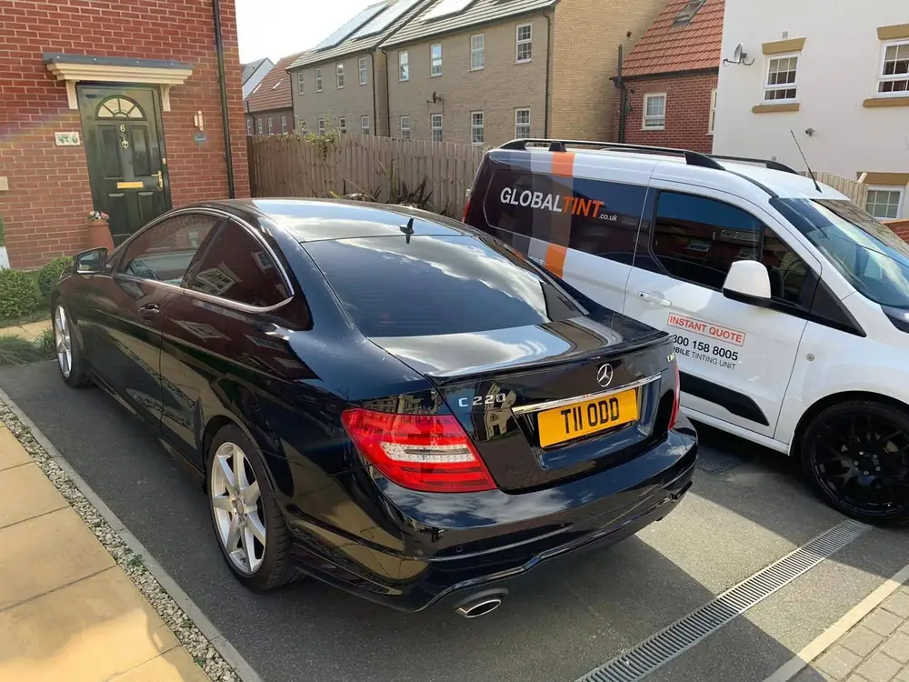 Black Mercedes-Benz C220 coupe with dark rear glass, parked on a driveway beside a Global Tint van