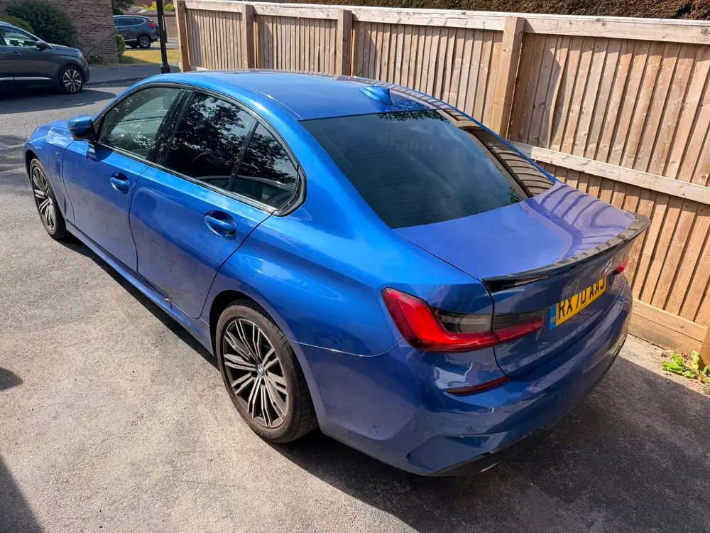 Blue BMW 3 Series with dark rear side windows and rear windscreen, parked beside a wooden fence