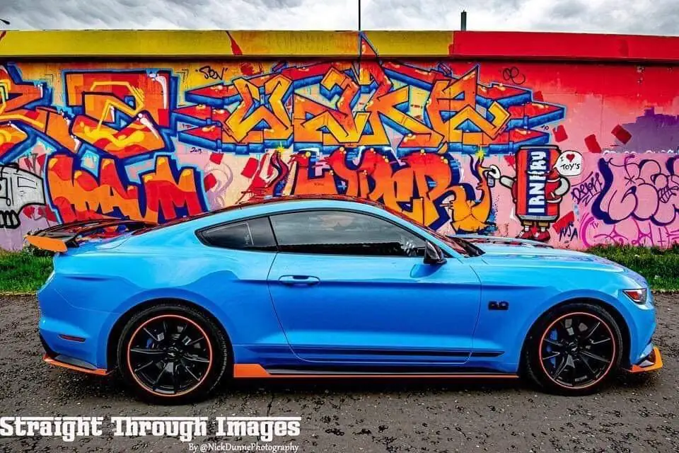 Blue coupe with orange trim and a dark rear side window, parked beside a wall covered in graffiti