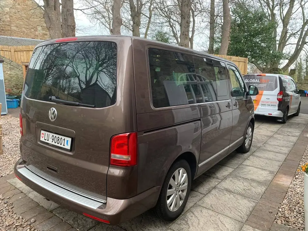 Brown van with dark rear side windows and rear window, parked on a paved driveway.