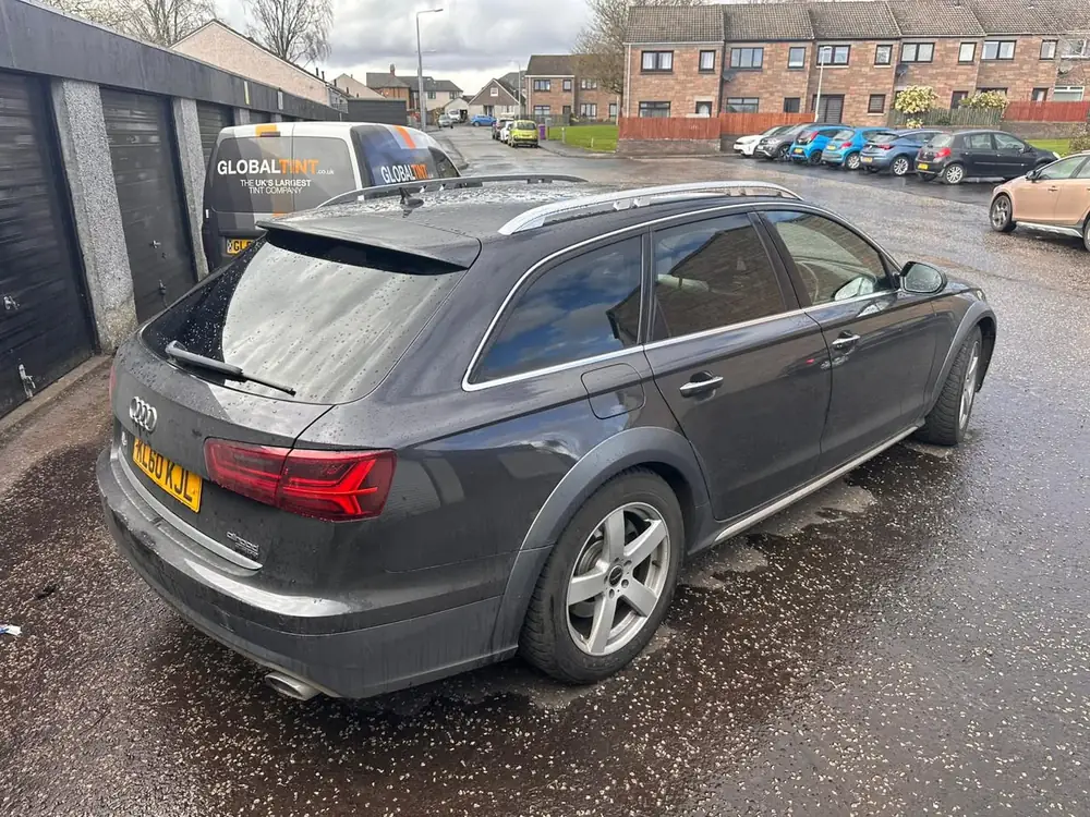 Dark grey Audi A6 Allroad with tinted rear glass, parked on wet ground beside garage doors