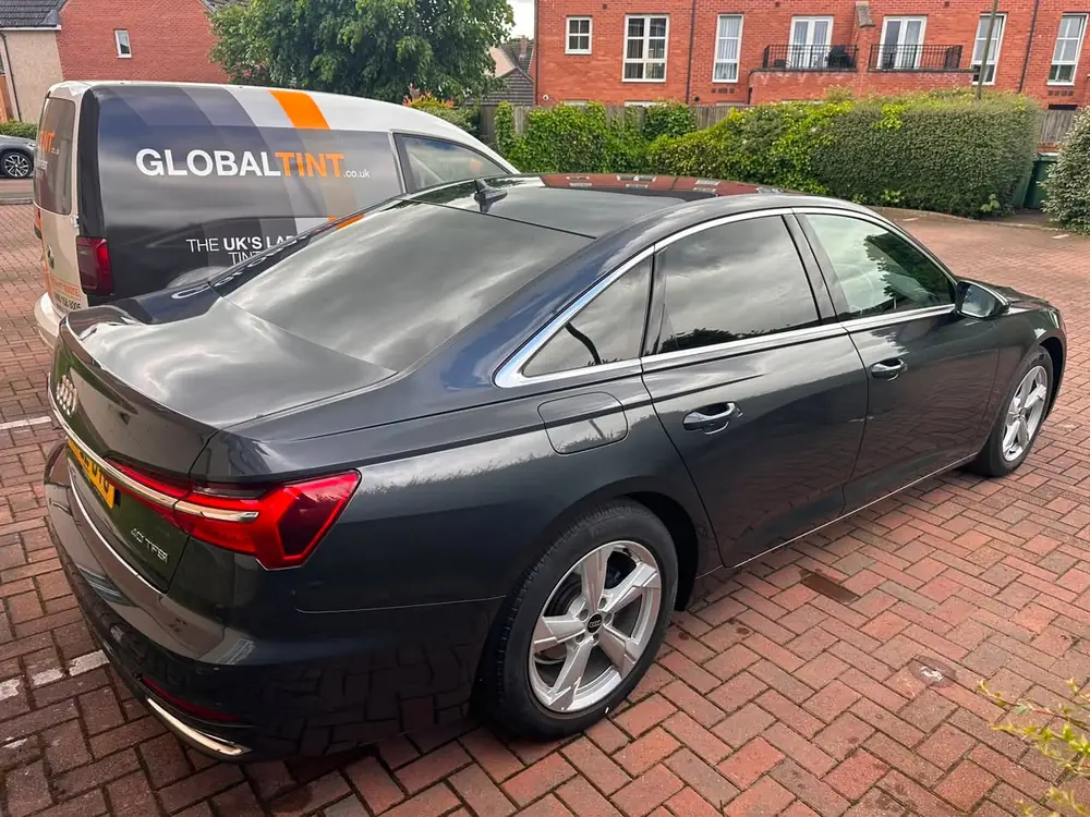 Dark grey Audi A6 with dark rear side windows, parked beside a Global Tint van on brick paving.