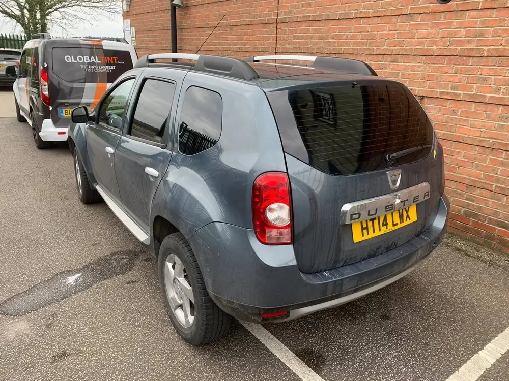 Grey Dacia Duster with dark rear side windows and rear windscreen, parked beside a brick wall.