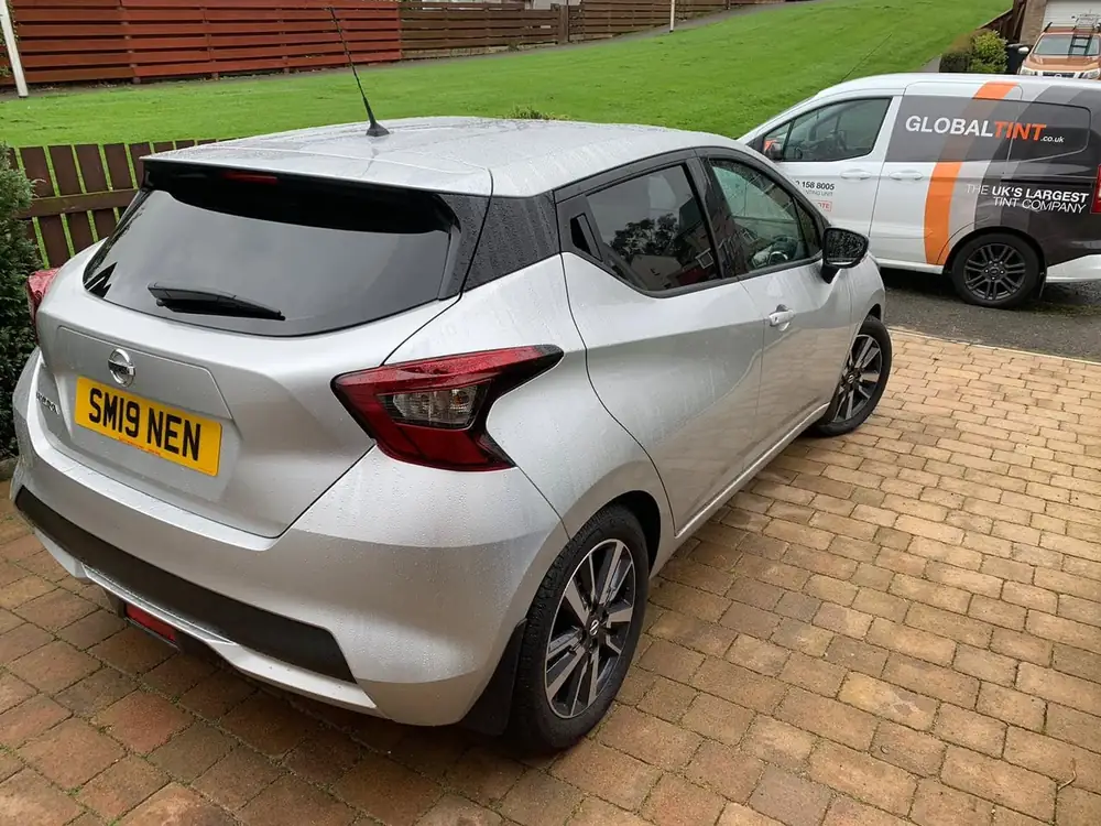 Silver Nissan Micra with a dark rear side window and rear windscreen, parked on paving beside a Global Tint van.