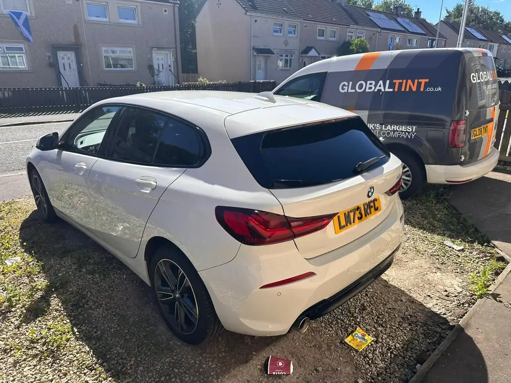 White BMW 1 Series with dark rear windows, parked on gravel beside a Global Tint van.