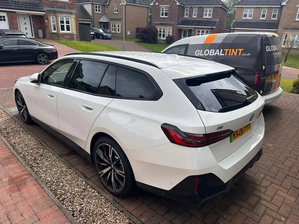 White BMW i5 Touring with dark rear side windows and rear windscreen, beside a Global Tint van on a driveway