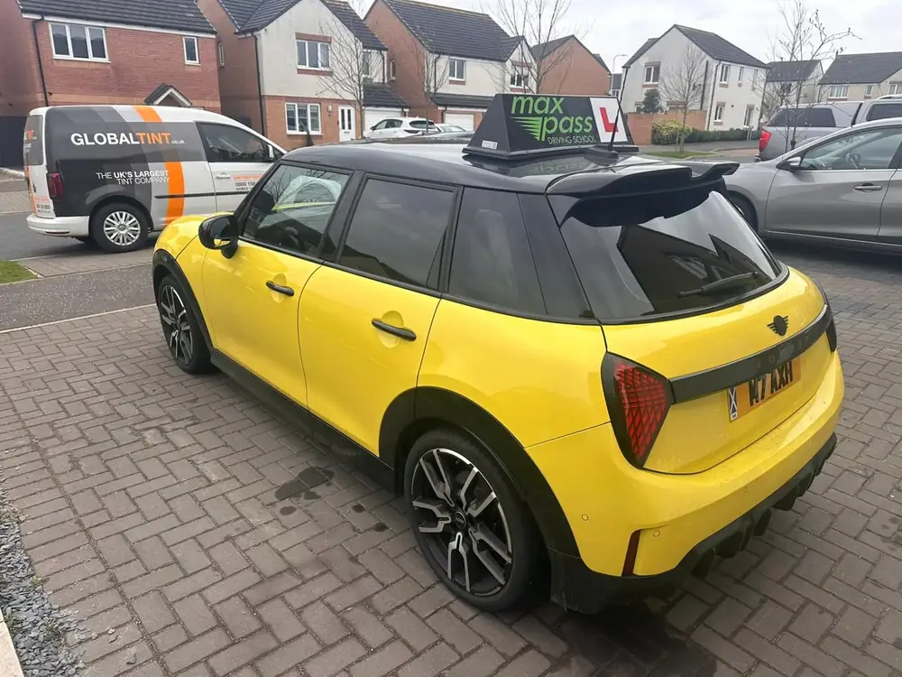 Yellow Mini Cooper with a black roof and dark tinted rear side windows and rear windscreen, parked on a paved driveway.